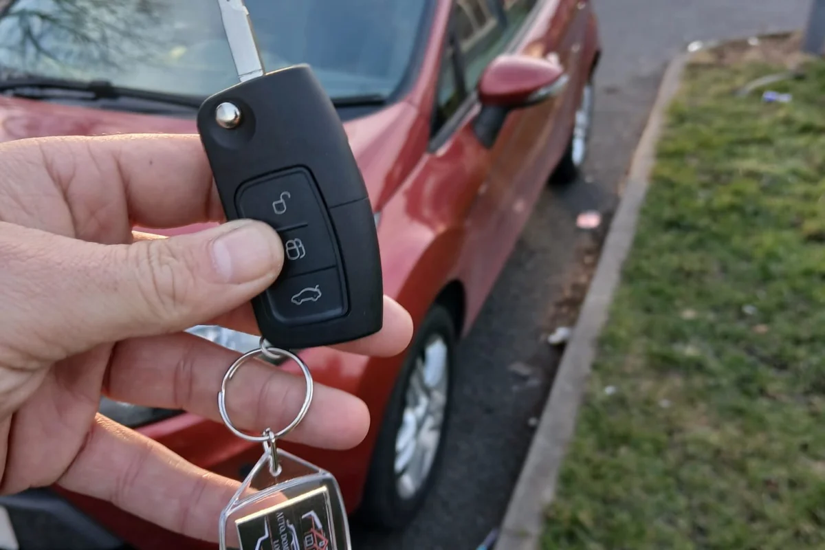 New car key and fob handed over beside a red hatchback